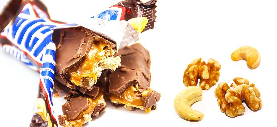 An image of opened Snicker showing the inside of the chocolate bar beside a couple of hazelnuts
